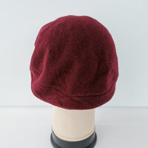May include: A burgundy-colored, wool newsboy cap. The cap is displayed on a mannequin head against a white background. The cap has a rounded crown and a small, stiff brim.