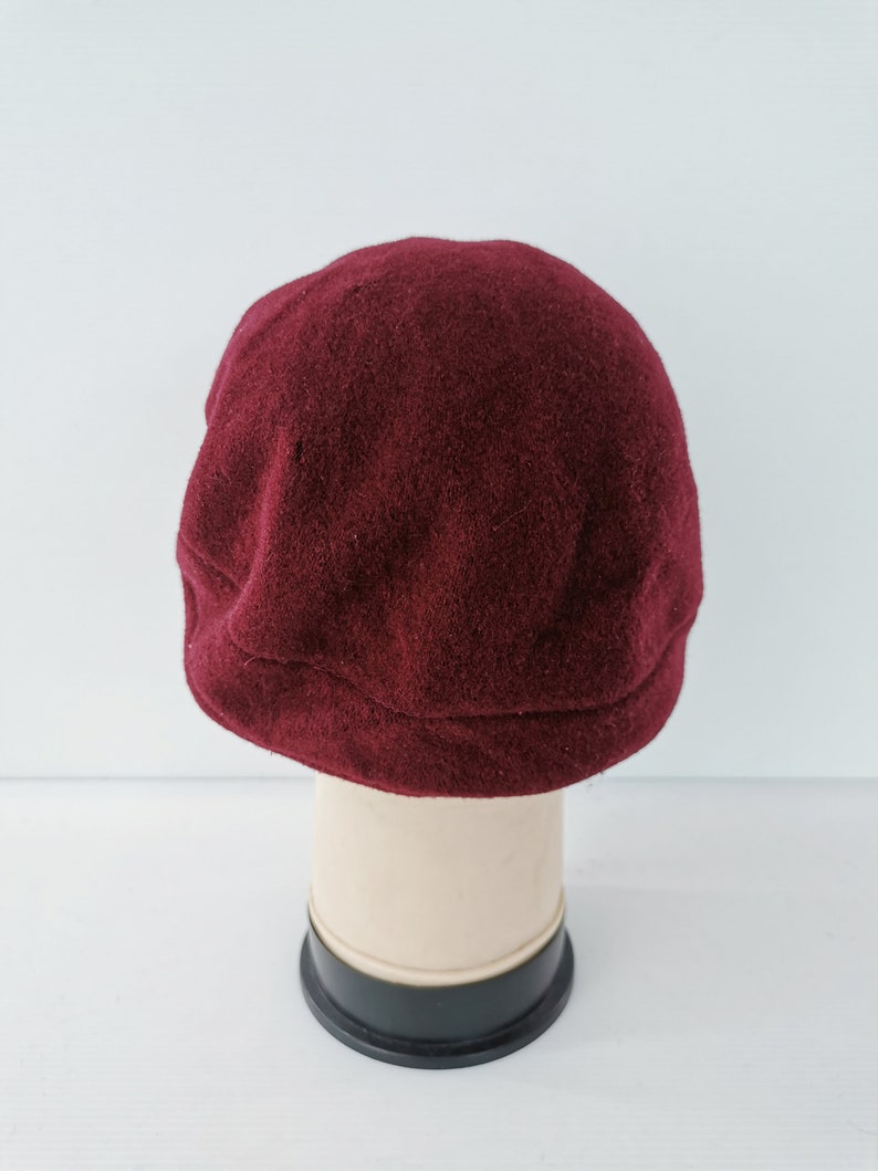 May include: A burgundy-colored, wool newsboy cap. The cap is displayed on a mannequin head against a white background. The cap has a rounded crown and a small, stiff brim.