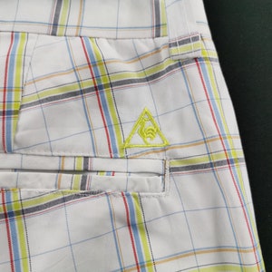 Op de afbeelding: Witte geruite shorts met een geel geborduurd driehoekig logo. Het logo toont een gestileerde vogel in de driehoek.