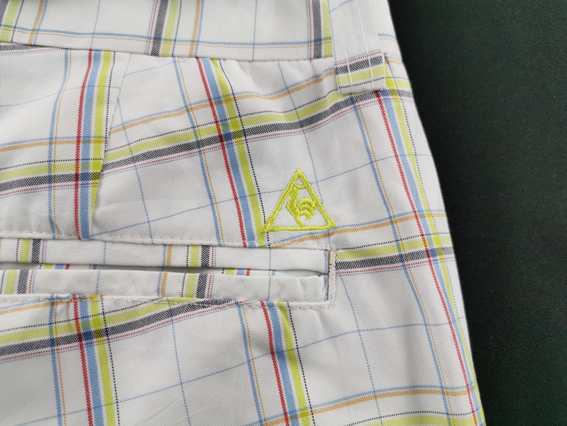 Op de afbeelding: Witte geruite shorts met een geel geborduurd driehoekig logo. Het logo toont een gestileerde vogel in de driehoek.
