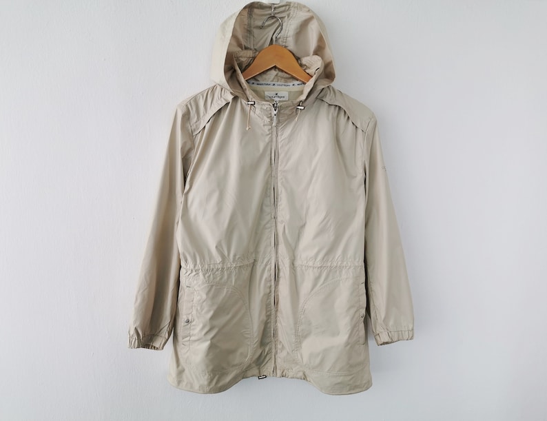May include: A beige hooded windbreaker jacket featuring a full-length front zipper, long sleeves with elasticized cuffs, and two front pockets. The jacket includes a drawstring hood with metal toggles and a cinched waist design for a structured fit.