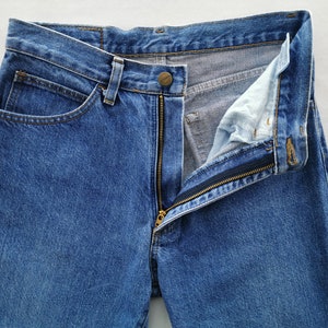 May include: Close-up of blue denim jeans with a partially unzipped brass zipper. The jeans have a button closure and classic five-pocket design. The fabric shows subtle wear and fading, typical of denim.