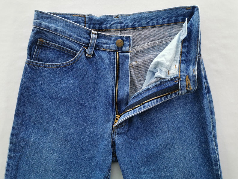 May include: Close-up of blue denim jeans with a partially unzipped brass zipper. The jeans have a button closure and classic five-pocket design. The fabric shows subtle wear and fading, typical of denim.