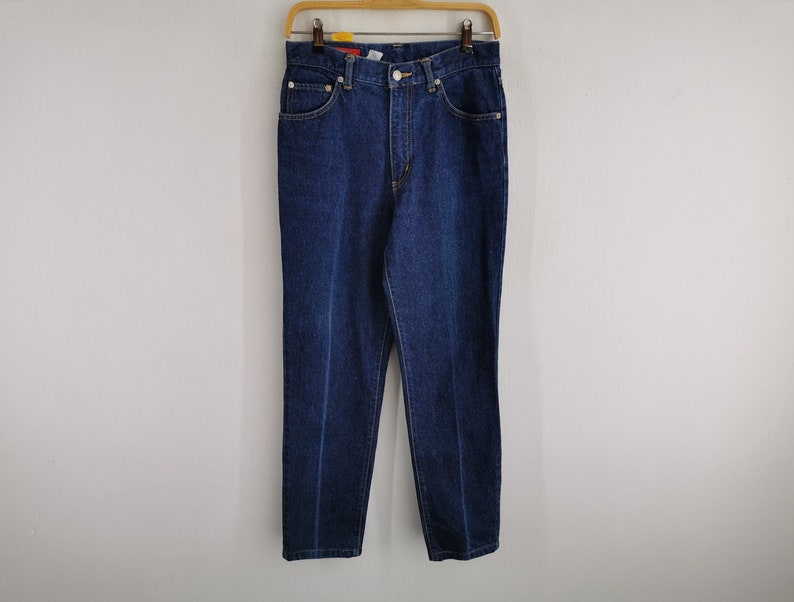 May include: Dark blue denim jeans hanging on a wooden hanger. The jeans have a classic five-pocket design with a button and zipper closure. The denim is a solid, medium-weight fabric.