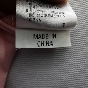 May include: A white fabric garment care label with black text reading MADE IN CHINA, positioned below a larger white tag containing Japanese text instructions regarding washing and tumble drying, attached to a light pink textile material.