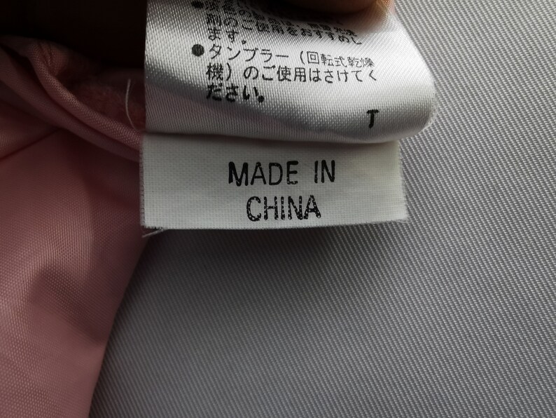 May include: A white fabric garment care label with black text reading MADE IN CHINA, positioned below a larger white tag containing Japanese text instructions regarding washing and tumble drying, attached to a light pink textile material.