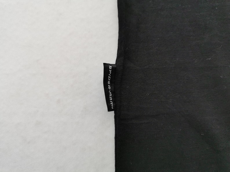 Pode incluir: Tecido preto com uma pequena etiqueta retangular anexada. A etiqueta &eacute; preta com texto branco que diz "SPIDER-MAN". O tecido &eacute; de cor preta s&oacute;lida e o fundo &eacute; branco.