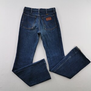 Peut inclure: Jean bleu fonc&eacute; avec une coupe &eacute;vas&eacute;e. Le jean pr&eacute;sente deux poches arri&egrave;re, un patch en cuir marron avec le nom de la marque et le logo Wrangler. Le jean est pos&eacute; &agrave; plat sur une surface blanche.
