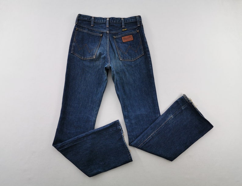 Peut inclure: Jean bleu fonc&eacute; avec une coupe &eacute;vas&eacute;e. Le jean pr&eacute;sente deux poches arri&egrave;re, un patch en cuir marron avec le nom de la marque et le logo Wrangler. Le jean est pos&eacute; &agrave; plat sur une surface blanche.
