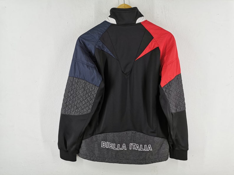 May include: A black, red, and blue athletic jacket with the text "BIELLA ITALIA" across the bottom. The jacket features a high collar and quilted gray panels on the sleeves. The design includes a red chevron on the back.
