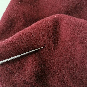 May include: Close-up of a burgundy fabric with a textured surface. A silver metal tool with a pointed tip is positioned near the fabric. The fabric appears to be a soft material, possibly wool or felt.