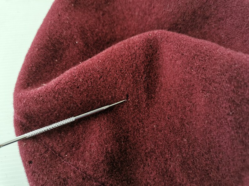 May include: Close-up of a burgundy fabric with a textured surface. A silver metal tool with a pointed tip is positioned near the fabric. The fabric appears to be a soft material, possibly wool or felt.