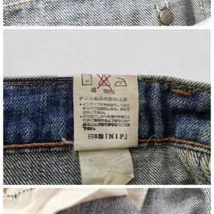 K&ouml;nnte beinhalten: Nahaufnahme von Jeans aus Denim mit mehreren Etiketten. Ein Etikett zeigt die Ma&szlig;e: Taille 76 cm, H&uuml;fte 93 cm und Gr&ouml;&szlig;e 30. Ein weiteres Etikett weist auf 100 % Baumwolle und Pflegehinweise hin. Ein drittes Etikett zeigt das Wrangler-Logo.