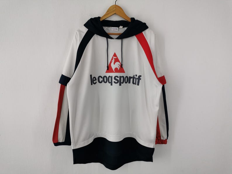 K&ouml;nnte beinhalten: Wei&szlig;er Kapuzenpullover mit schwarzen und roten Akzenten. Das Le Coq Sportif-Logo ist in Rot und Schwarz dargestellt. Er hat eine Kapuze mit Kordelzug und lange &Auml;rmel. Der Saum und die Manschetten sind schwarz. Ein sportliches und l&auml;ssiges Kleidungsst&uuml;ck.
