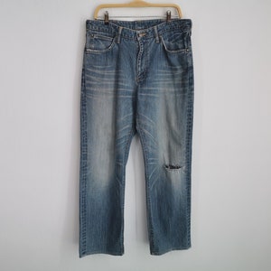 May include: Faded blue denim jeans with a distressed look, featuring a worn appearance and a tear on the right knee. The jeans are hanging on a wooden hanger against a plain white wall.