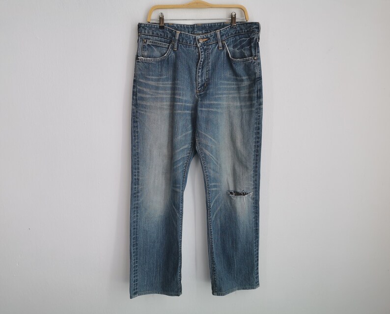 May include: Faded blue denim jeans with a distressed look, featuring a worn appearance and a tear on the right knee. The jeans are hanging on a wooden hanger against a plain white wall.
