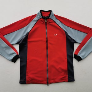 Op de afbeelding: Een rode, grijze en zwarte trainingsjas met ritssluiting en het Nike-logo. De jas heeft een rode body, grijze mouwen en zwarte accenten. De Nike swoosh is wit.