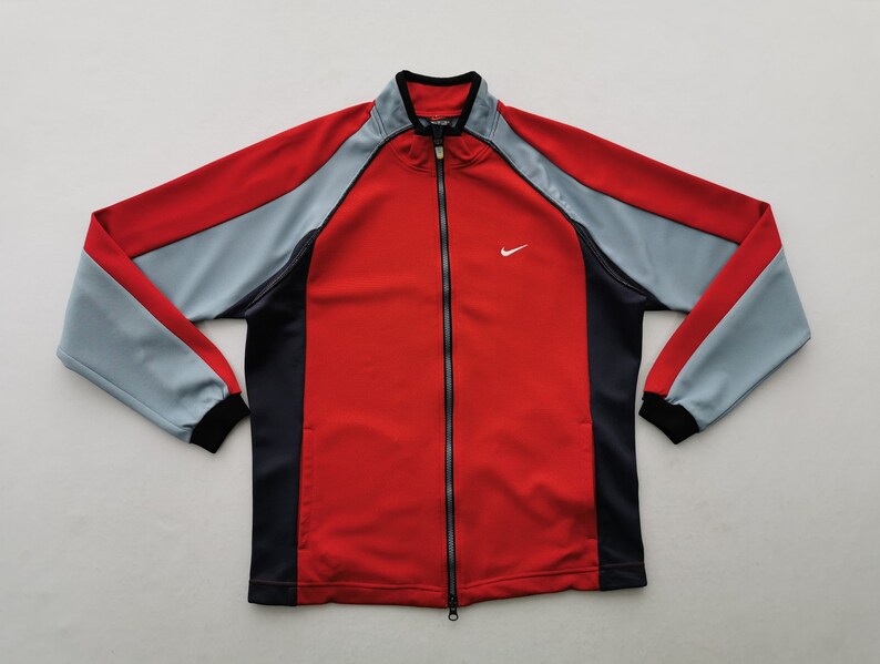 Op de afbeelding: Een rode, grijze en zwarte trainingsjas met ritssluiting en het Nike-logo. De jas heeft een rode body, grijze mouwen en zwarte accenten. De Nike swoosh is wit.