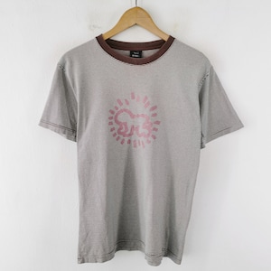 Pode incluir: Uma camiseta listrada cinza e branca com gola e mangas marrons. A camisa apresenta um gr&aacute;fico rosa na frente. A camiseta est&aacute; pendurada em um cabide de madeira contra uma parede branca.