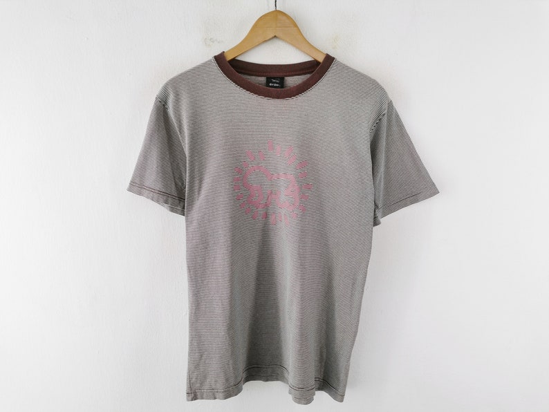 Pode incluir: Uma camiseta listrada cinza e branca com gola e mangas marrons. A camisa apresenta um gr&aacute;fico rosa na frente. A camiseta est&aacute; pendurada em um cabide de madeira contra uma parede branca.
