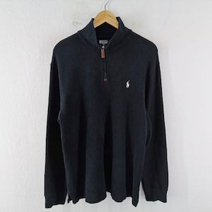 Puede incluir: Un jersey de manga larga azul marino con cierre de cremallera de un cuarto y un pequeño logotipo blanco en el pecho. El jersey tiene cuello y puños acanalados. El tirador de la cremallera es marrón.