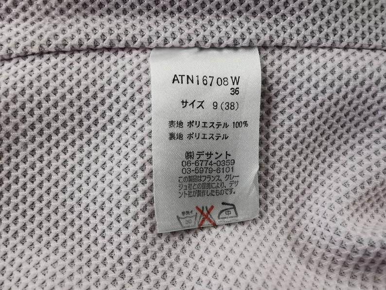 Puede incluir: Primer plano de una etiqueta blanca con texto japon&eacute;s y los n&uacute;meros "ATN16708 W" y "36". La etiqueta tambi&eacute;n incluye la talla "9 (38)" e instrucciones de lavado. La etiqueta est&aacute; sobre una tela texturizada de color lila claro.