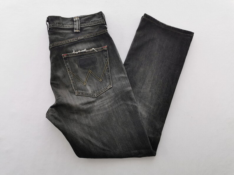 May include: A pair of black denim jeans with a faded wash. The jeans feature a classic five-pocket design, a button and zipper closure, and a leather patch on the back pocket. The back pocket has a yellow stitched "W" design.
