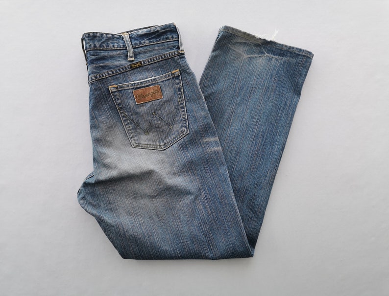 May include: Blue denim Wrangler jeans with a faded wash. The jeans have a classic five-pocket design, a brown leather patch with the Wrangler logo, and a straight leg. The jeans are folded and laid flat on a white surface.