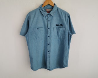 Puede incluir: Una camisa de manga corta de color azul claro con cuello y dos bolsillos en el pecho. La palabra XLARGE está bordada en hilo negro sobre el bolsillo izquierdo. La camisa cuenta con una tapeta central de botones y un colgador de madera.