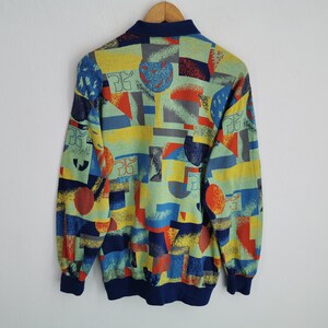 May include: A vintage sweatshirt with a colorful geometric pattern. The design features shapes in blue, red, yellow, and gray on a light green background. It has a navy blue collar and cuffs, and is displayed on a wooden hanger.