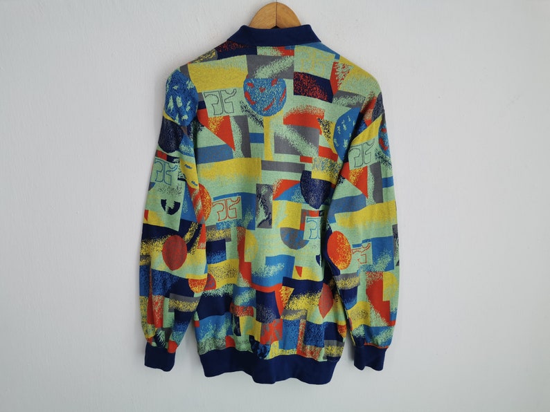 May include: A vintage sweatshirt with a colorful geometric pattern. The design features shapes in blue, red, yellow, and gray on a light green background. It has a navy blue collar and cuffs, and is displayed on a wooden hanger.
