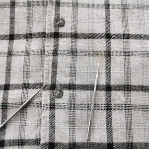 May include: Two silver metal tools with pointed ends rest on a gray and black plaid fabric. The tools appear to be small and slender, possibly for detailed work or crafts. The fabric has a buttoned closure.