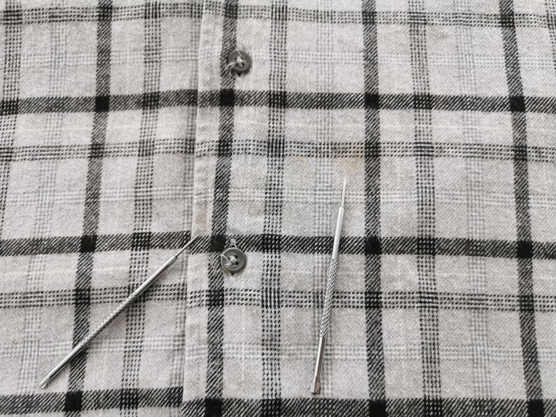 May include: Two silver metal tools with pointed ends rest on a gray and black plaid fabric. The tools appear to be small and slender, possibly for detailed work or crafts. The fabric has a buttoned closure.