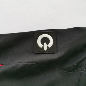 Pode incluir: Tecido preto com um patch de logotipo branco. O patch &eacute; quadrado com um design branco estilizado. Uma faixa vermelha &eacute; vis&iacute;vel no lado esquerdo da imagem. O tecido parece fazer parte de uma pe&ccedil;a de vestu&aacute;rio.