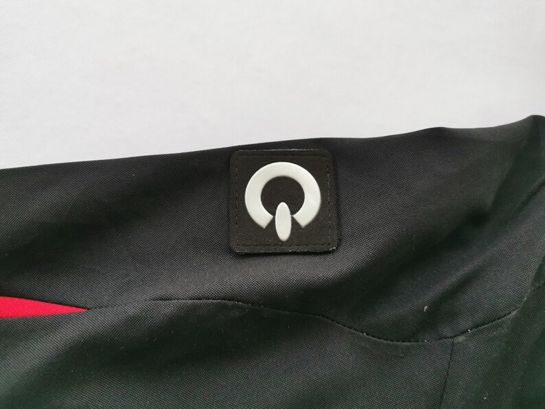 Pode incluir: Tecido preto com um patch de logotipo branco. O patch &eacute; quadrado com um design branco estilizado. Uma faixa vermelha &eacute; vis&iacute;vel no lado esquerdo da imagem. O tecido parece fazer parte de uma pe&ccedil;a de vestu&aacute;rio.