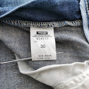 May include: Close-up of blue denim jeans with a white label. The label says "Wrangler" and the size is "30". The jeans have visible stitching and a light blue and white striped detail. The jeans are made of denim.