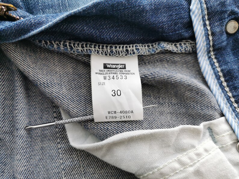 May include: Close-up of blue denim jeans with a white label. The label says "Wrangler" and the size is "30". The jeans have visible stitching and a light blue and white striped detail. The jeans are made of denim.