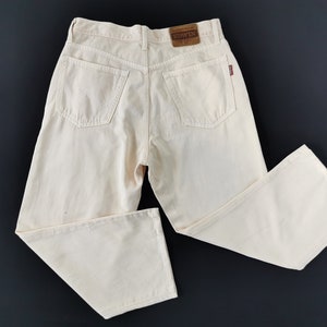 Può includere: Jeans in denim color crema con un classico design a cinque tasche. I jeans presentano una toppa in pelle marrone con il nome del marchio e una piccola etichetta rossa sulla tasca posteriore. I pantaloni sono appoggiati su una superficie scura.