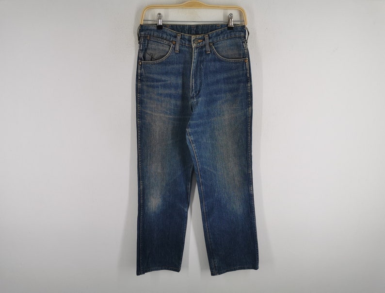 K&ouml;nnte beinhalten: Blaue Jeans aus Denim, h&auml;ngen an einem Holzkleiderb&uuml;gel. Die Jeans haben ein klassisches F&uuml;nf-Taschen-Design, einen Knopf- und Rei&szlig;verschluss und eine gerade Beinpassform. Der Denim hat ein verwaschenes, abgenutztes Aussehen.