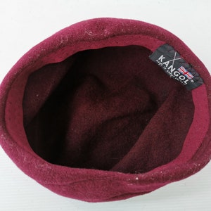 May include: A burgundy Kangol hat, viewed from the inside. The hat is made of a soft, felt-like material. The interior features a dark burgundy lining. A black tag with the Kangol logo and a British flag is visible.