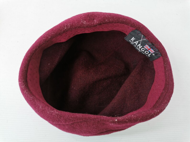 May include: A burgundy Kangol hat, viewed from the inside. The hat is made of a soft, felt-like material. The interior features a dark burgundy lining. A black tag with the Kangol logo and a British flag is visible.