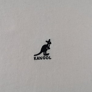 Peut inclure: Gros plan sur un tissu beige clair avec un logo kangourou noir et le mot "KANGOL" imprim&eacute; en dessous. Le logo est une silhouette de kangourou. Le tissu a une texture subtile.