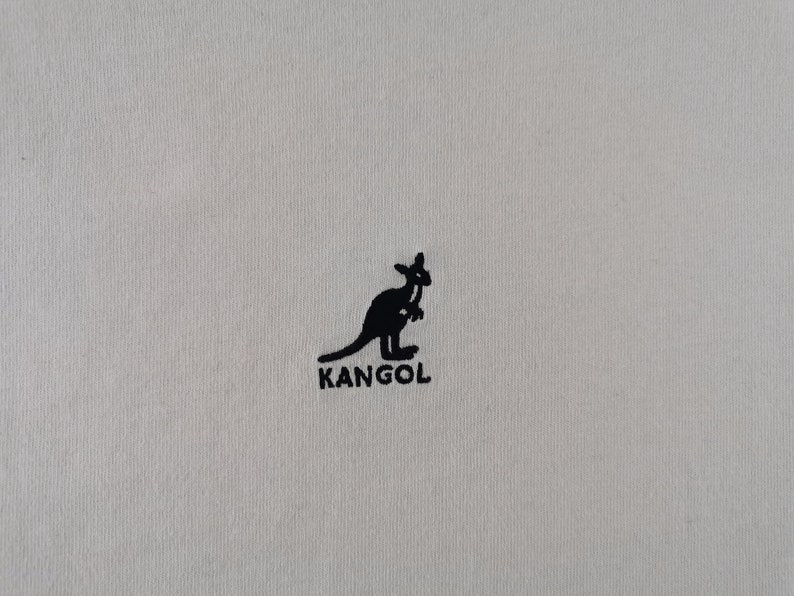 Peut inclure: Gros plan sur un tissu beige clair avec un logo kangourou noir et le mot "KANGOL" imprim&eacute; en dessous. Le logo est une silhouette de kangourou. Le tissu a une texture subtile.