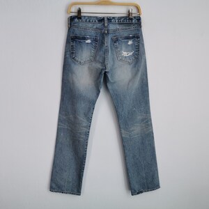 May include: Light blue denim jeans with distressed details, including rips and fading. The jeans are displayed on a wooden hanger against a white background. The back pockets and belt loops are visible.