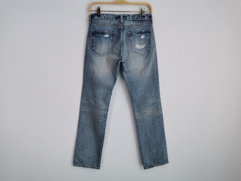 May include: Light blue denim jeans with distressed details, including rips and fading. The jeans are displayed on a wooden hanger against a white background. The back pockets and belt loops are visible.