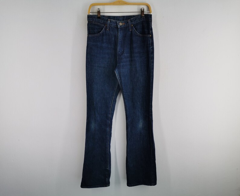 Peut inclure: Un jean en denim bleu fonc&eacute; avec une conception classique &agrave; cinq poches, suspendu &agrave; un cintre en bois. Le jean a un l&eacute;ger &eacute;vasement en bas et montre une certaine usure. Le jean est de couleur moyenne et semble vintage.