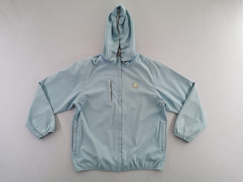 May include: A light blue hooded jacket with a zipper closure and two front pockets. The jacket has a small square emblem on the chest and a hood. The sleeves have elastic cuffs. This lightweight jacket is suitable for outdoor activities.