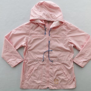 May include: A light pink hooded jacket featuring a full-length front zipper, long sleeves, and two front pockets. The garment includes blue drawstring cords with silver-toned toggles at the hood and waist, and a small square logo patch.