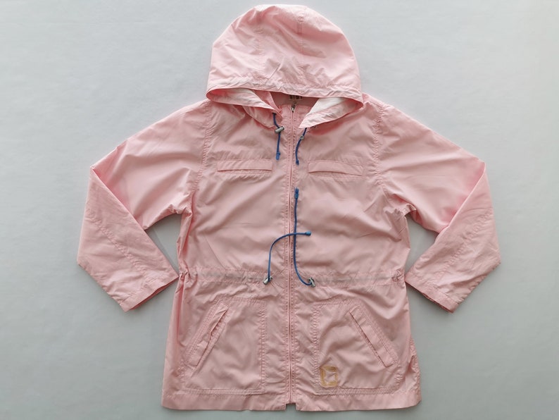 May include: A light pink hooded jacket featuring a full-length front zipper, long sleeves, and two front pockets. The garment includes blue drawstring cords with silver-toned toggles at the hood and waist, and a small square logo patch.
