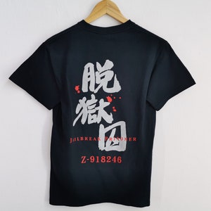 May include: A black short-sleeved t-shirt featuring large grey Japanese kanji characters on the back. Below the characters, the red text reads JAELBREAK PRISONER and Z-918246. Small red paint-like splatters are visible around the grey text.
